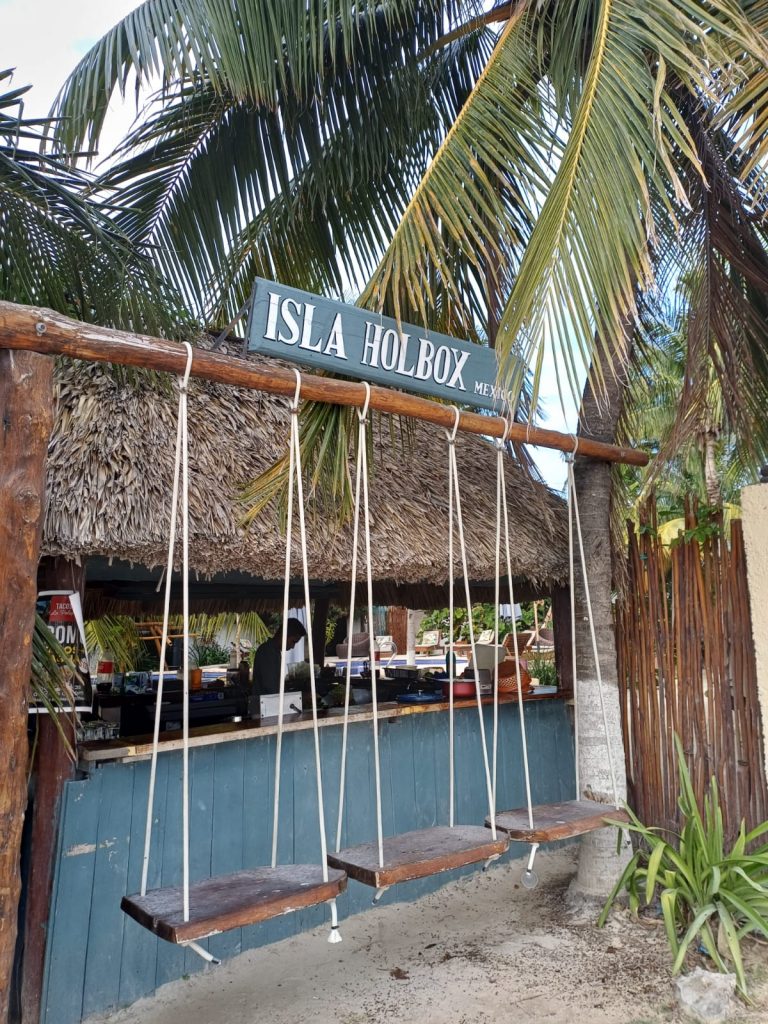 Columpio de madera en la playa con el cartel superior que dice “Isla de Holbox”, frente al mar turquesa