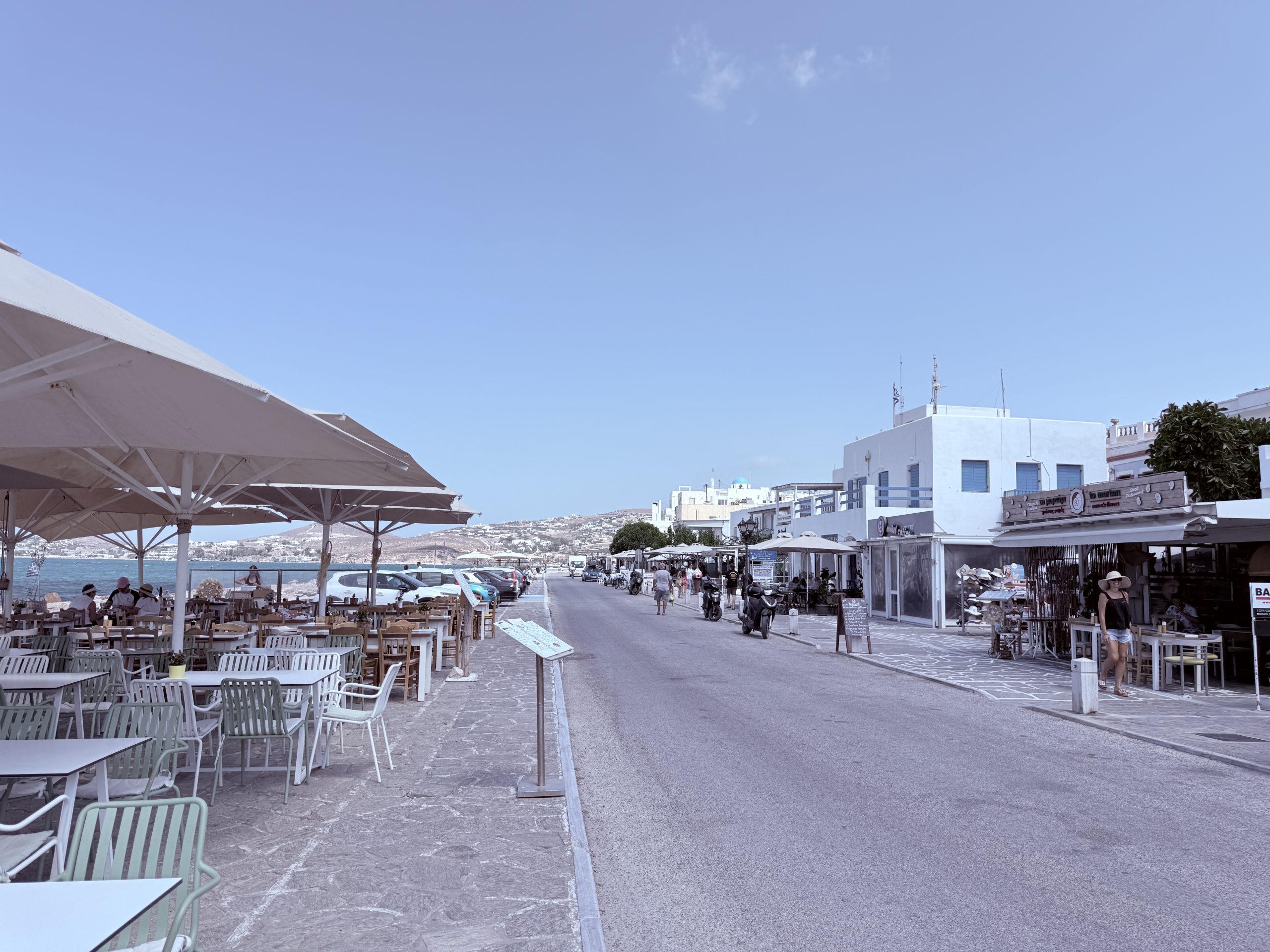 Vista de la carretera principal junto al mar en Parikia, Paros, con terrazas de restaurante, casas blancas y el mar Egeo al fondo