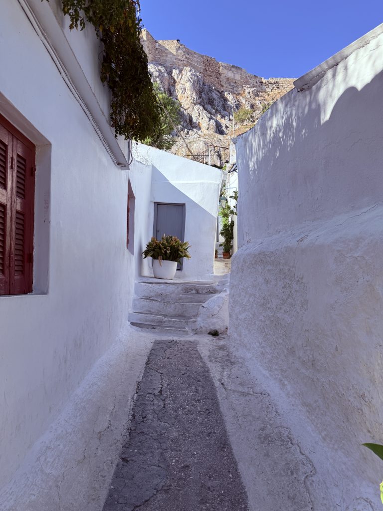 Calle estrecha y blanca del barrio de Anafiotika en Atenas, con plantas y casas tradicionales, y la Acrópolis al fondo