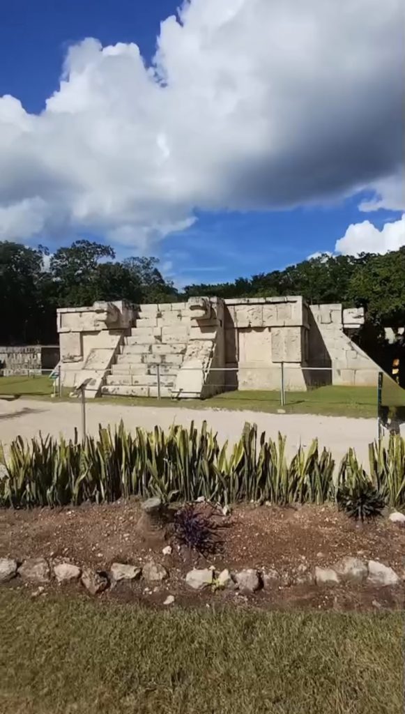 Vista del Templo del Hombre Barbado en Chichén Itzá, con escalinatas y cabezas de serpiente talladas, bajo un cielo azul con nubes