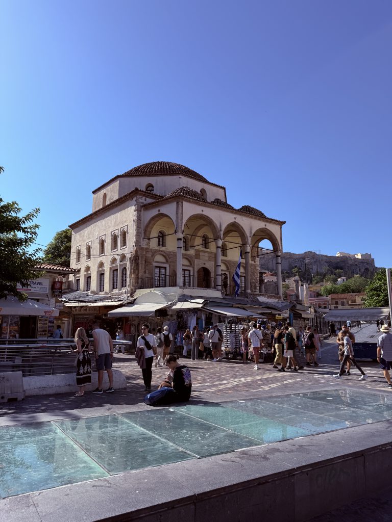 Plaza Monastiraki en Atenas con puestos, gente paseando, y la Acrópolis visible al fondo en una tarde soleada